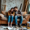 Ремонт в квартире - девушка и парень очень уставшие сидят на диване, вокруг валяются обрывки обоев, инструмент